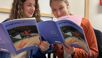 Zwei lächelnde Frauen sitzen zusammen und lesen Broschüren der jungen Caritas, die Informationen zur Natur enthalten.