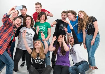 Gruppe junger Menschen, die fröhlich in die Kamera lächeln und hoch die Hände halten, während sie ein Selfie machen