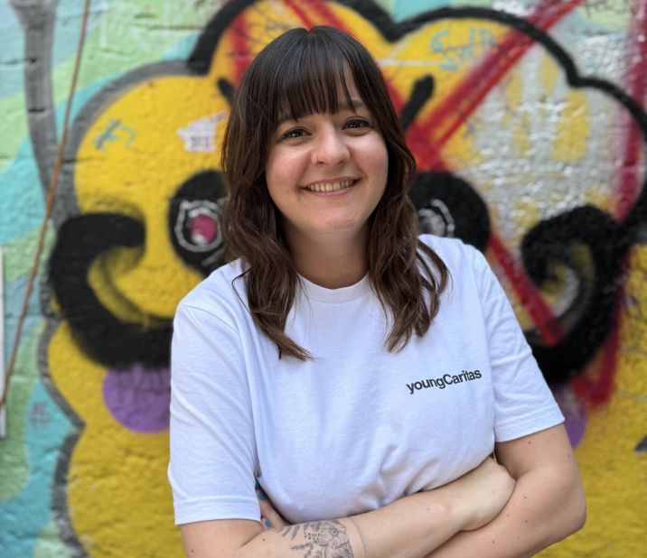Eine junge Frau mit lächelndem Gesicht steht vor einer farbenfrohen Graffiti-Wand. Sie trägt ein weißes T-Shirt mit dem Schriftzug 'youngCaritas'.