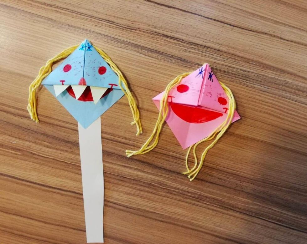 Zwei bunte Papiermonster mit verschiedenen Gesichtsausdrücken, einer hat blaue und der andere rosa Farben, beide mit Haaren aus Wolle.