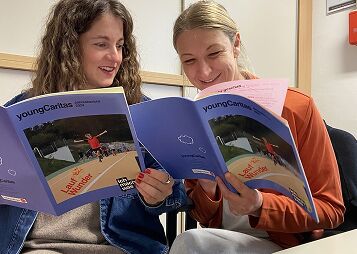 Zwei lächelnde Frauen sitzen zusammen und lesen Broschüren der jungen Caritas, die Informationen zur Natur enthalten.