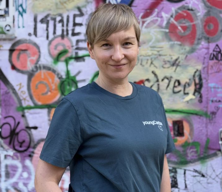 Eine Person steht vor einer bunten Graffiti-Wand und lächelt. Sie trägt ein dunkelgrünes T-Shirt und Jeans.