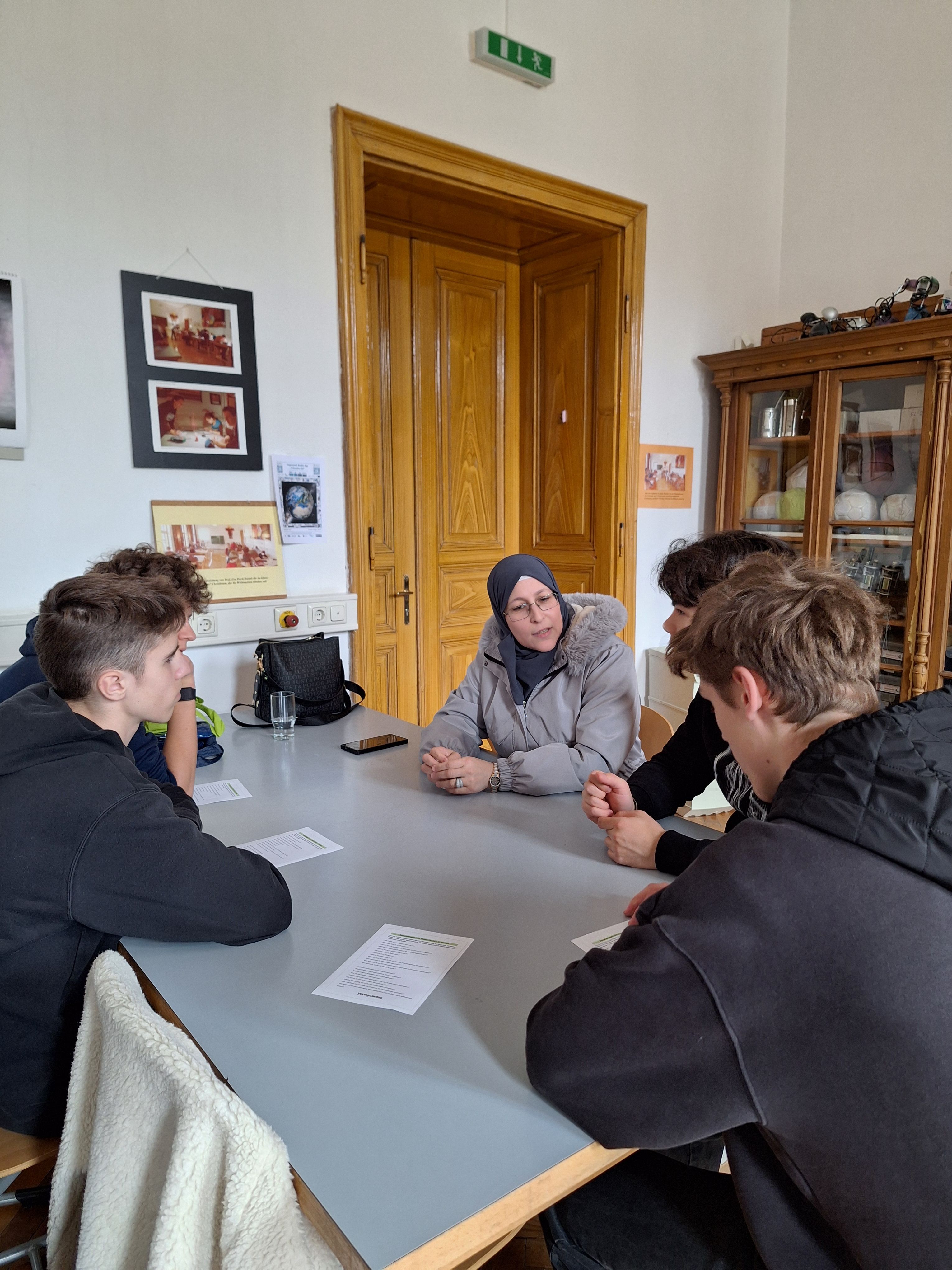 Vier Jugendliche sitzen an einem Tisch und diskutieren mit einer älteren Frau in einem hellen Raum.