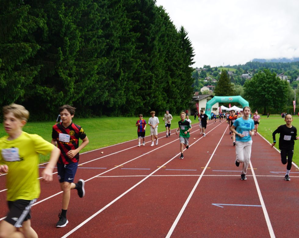 Eine Gruppe von Schülern läuft auf einer Laufbahn während eines Wettbewerbs, umgeben von Bäumen und einer Sporteinrichtung.
