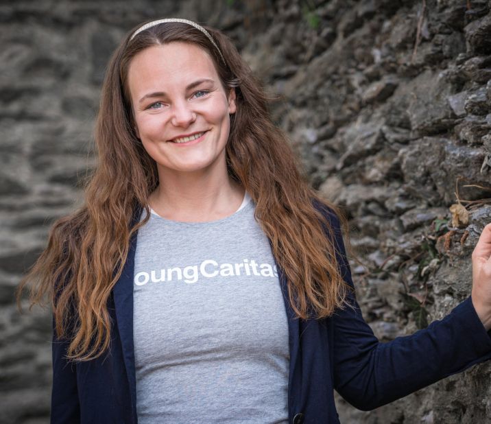 Eine junge Frau mit langen Haaren lächelt vor einer Steinmauer. Sie trägt ein graues Shirt mit der Aufschrift 'YoungCaritas'.