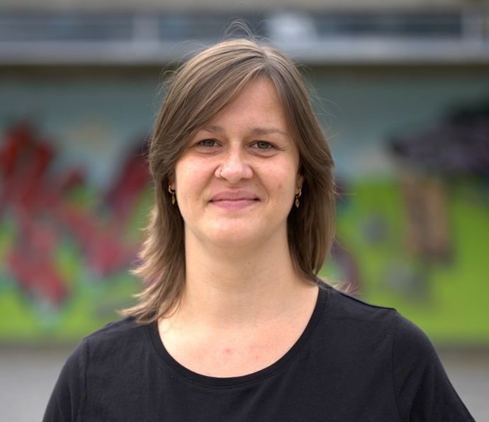 Frau mit mittellangen Haaren lächelt in die Kamera, trägt ein schwarzes T-Shirt mit der Aufschrift youngCaritas. Der Hintergrund ist bunt.