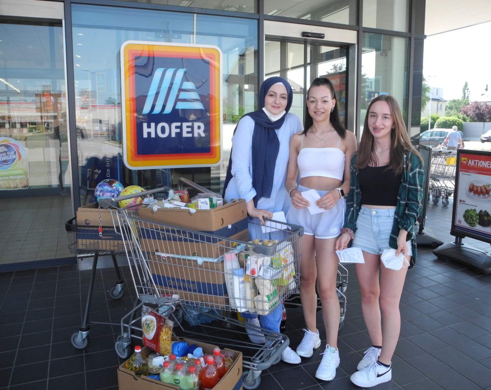 Drei junge Frauen stehen vor dem Hofer-Supermarkt mit einem voll beladenen Einkaufswagen. Es ist ein sonniger Tag.