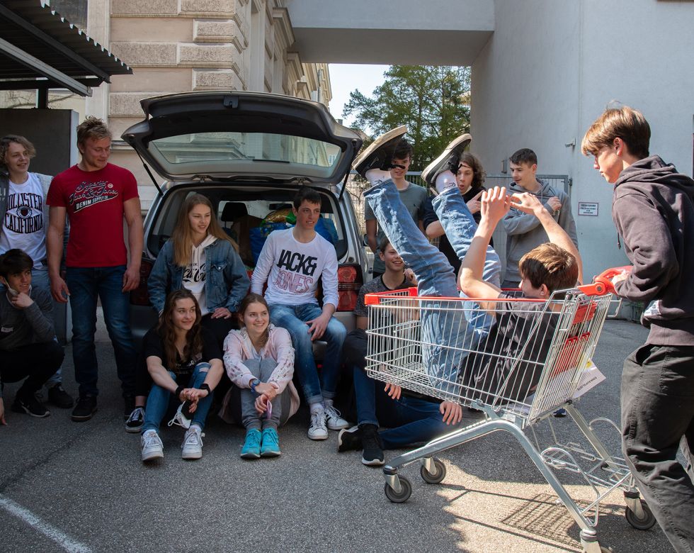 Gruppe von Jugendlichen vor einem geöffneten Auto auf einem Innenhof; ein Jugendlicher liegt lachend kopfüber in einem Einkaufswagen, während andere danebenstehen oder sitzen und die Szene beobachten