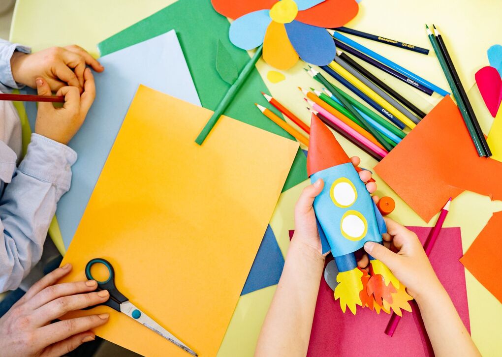 Zwei Kinderhände arbeiten mit buntem Papier, Scheren und Stiften an Bastelprojekten, darunter eine selbstgemachte Rakete.
