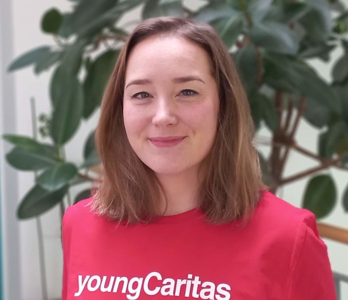 Eine junge Frau in einem roten T-Shirt mit dem Logo youngCaritas, steht vor einer Pflanze und lächelt.