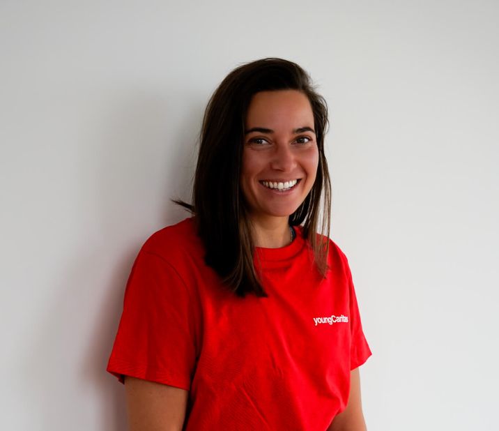 Eine junge Frau mit langen, dunklen Haaren trägt ein rotes T-Shirt und Jeans. Sie lächelt fröhlich und steht an einer weißen Wand.