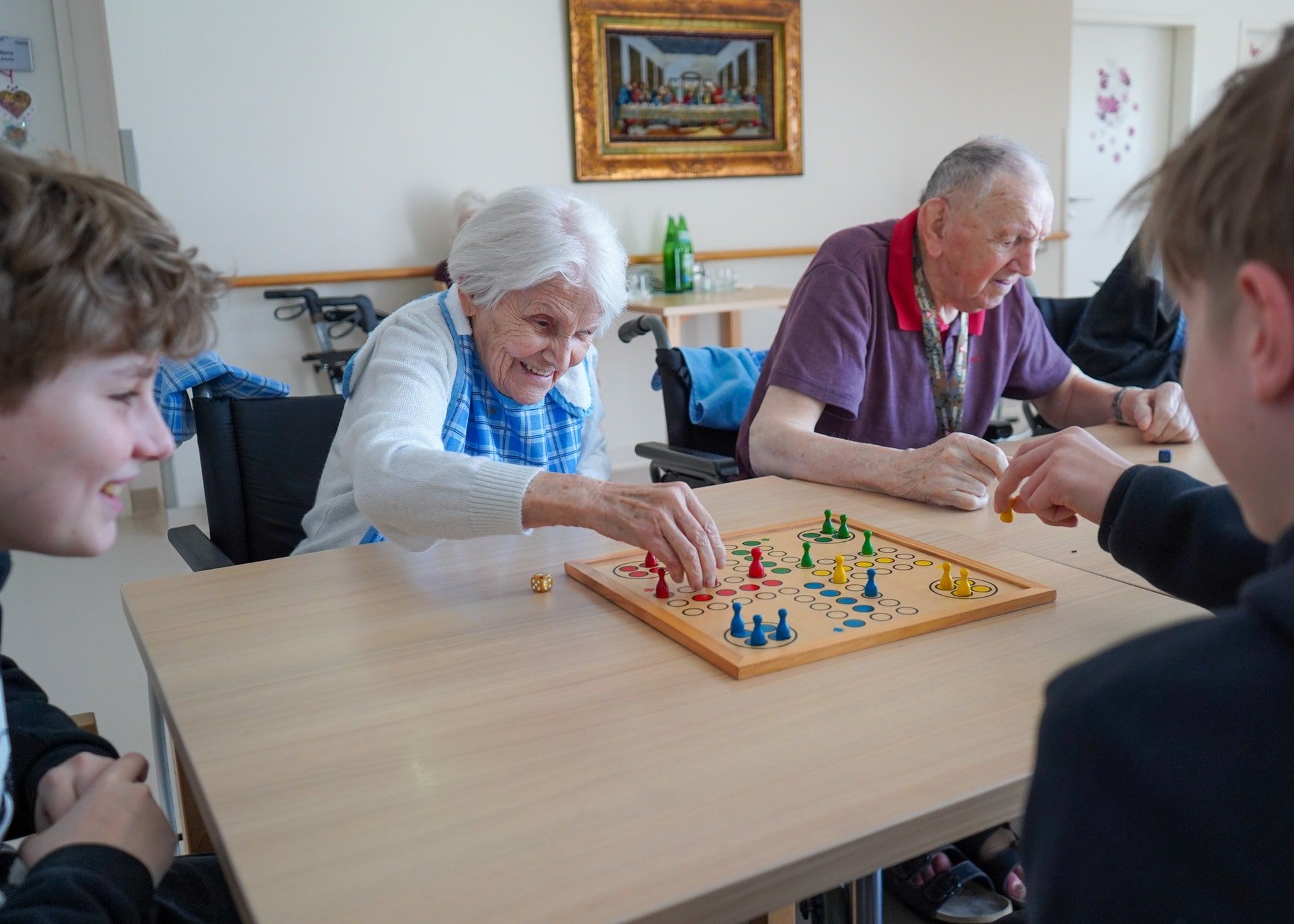 Ältere Menschen spielen ein Brettspiel mit jungen Besuchern in einer Betreuungseinrichtung. Lachen und Interaktion fördern die Gemeinschaft.