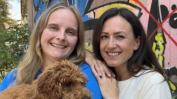 Zwei lächelnde Frauen, eine hält einen braunen Hund, bunte Graffiti im Hintergrund