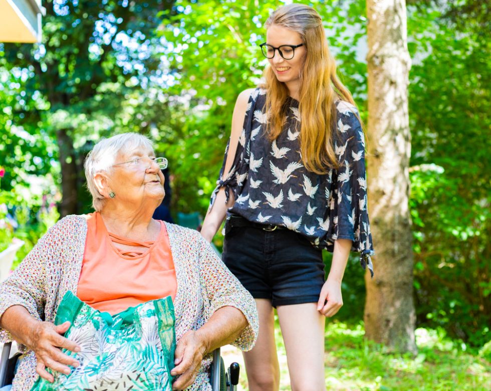 Eine junge Frau steht neben einer älteren Dame im Rollstuhl.