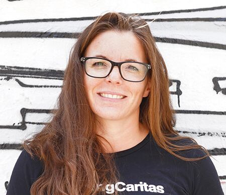 Eine lächelnde Frau mit langen Haaren trägt ein schwarzes T-Shirt mit der Aufschrift 'gCaritas' vor einer grauen Wand.