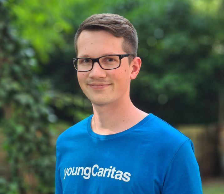 Ein junger Mann trägt ein blaues T-Shirt mit der Aufschrift youngCaritas und lächelt in eine grüne Umgebung.