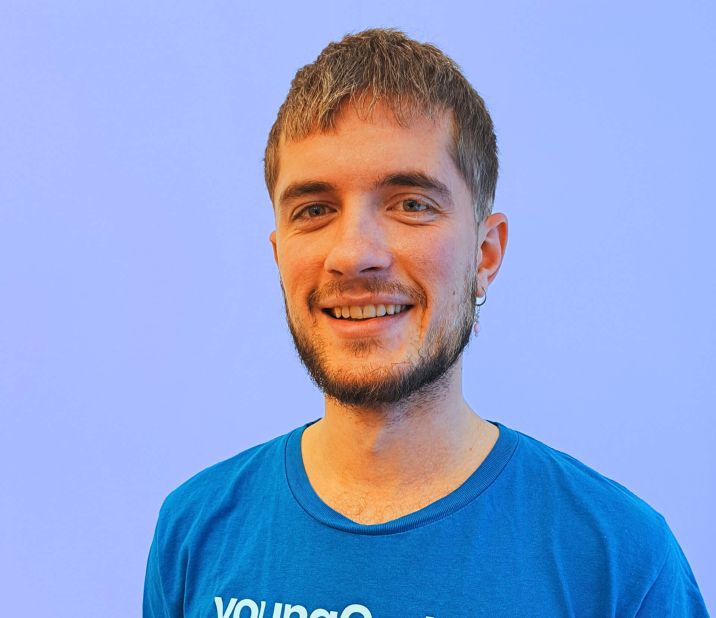 Ein junger Mann lächelt mit einem blauen T-Shirt von youngCaritas vor einem hellblauen Hintergrund.