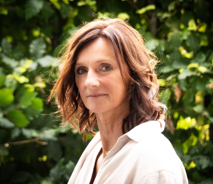 Eine Frau mit lockigem Haar steht vor einem grünen Hintergrund und blickt nachdenklich in die Kamera.