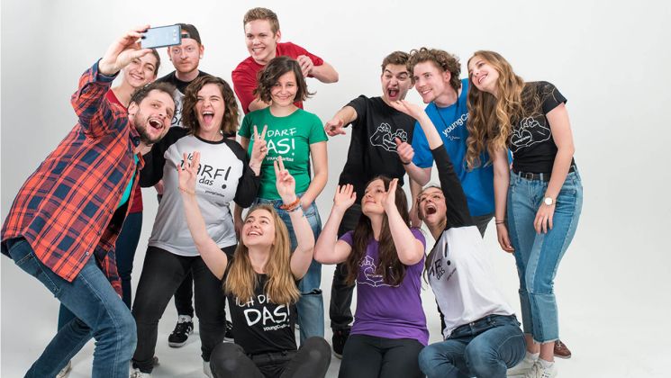Gruppe junger Menschen, die fröhlich in die Kamera lächeln und hoch die Hände halten, während sie ein Selfie machen