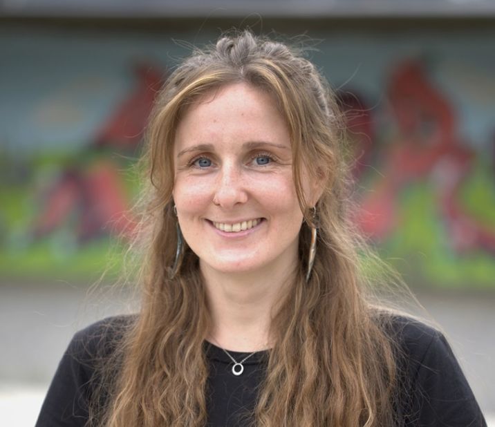 Eine junge Frau mit langen, lockigen Haaren und einem freundlichen Lächeln, die vor einem bunten Wandgemälde steht.
