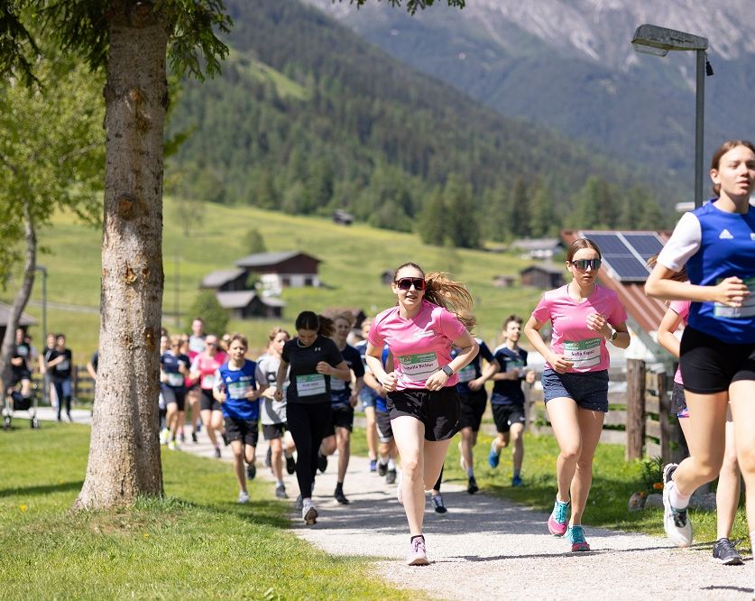 Eine Gruppe von Läufern, darunter Frauen in pinken T-Shirts, läuft auf einem Kiesweg mit Bergen im Hintergrund.
