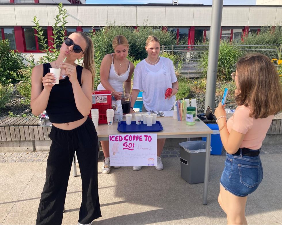 Eine Gruppe von jungen Frauen verkauft Iced Coffee. Eine steht vorne mit einem Getränk, die anderen helfen am Stand.