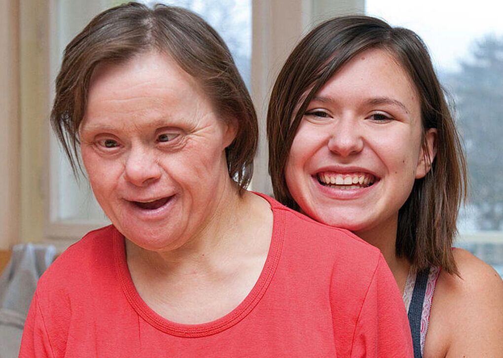 Zwei Frauen lächeln zusammen, eine sitzt vorne mit Down-Syndrom und die andere steht lächelnd dahinter.