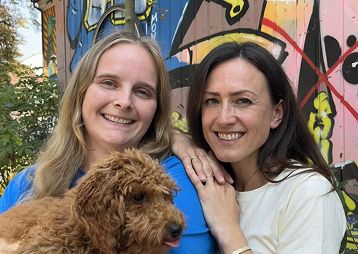Zwei lächelnde Frauen, eine hält einen braunen Hund, bunte Graffiti im Hintergrund