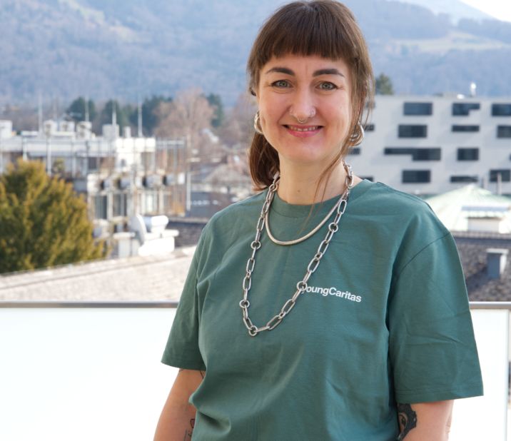 Eine Frau mit kurzer brauner Frisur steht auf einem Balkon. Sie trägt ein grünes T-Shirt und eine Kette, mit Bergen im Hintergrund.