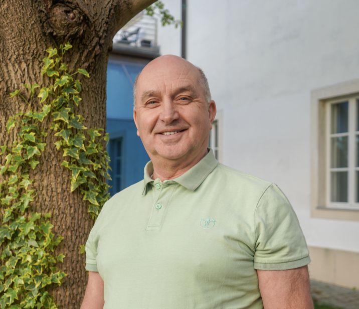 Ein älterer Mann steht lächelnd vor einem Baum mit Efeu, in einem grünen Poloshirt, mit einem Gebäude im Hintergrund.