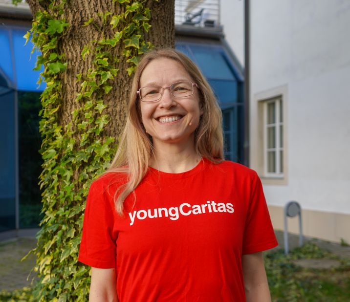 Die junge Frau lächelt vor einem Baum und trägt ein rotes T-Shirt mit der Aufschrift youngCaritas.