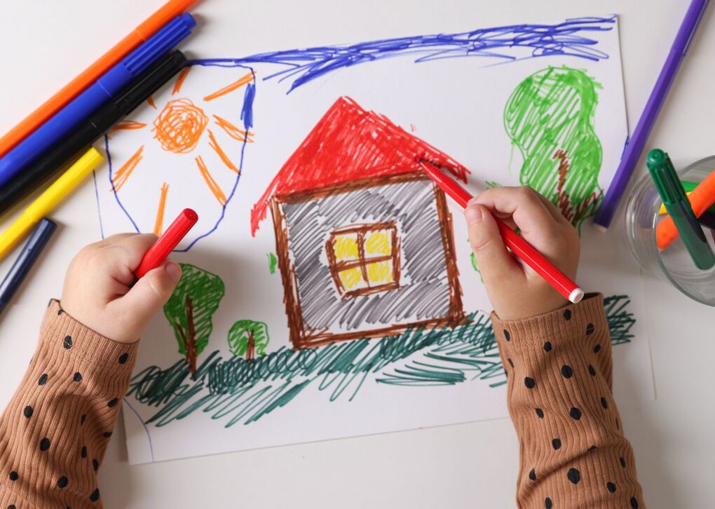 Ein Kind zeichnet ein farbenfrohes Haus mit verschiedenen Markern auf einem Blatt Papier.