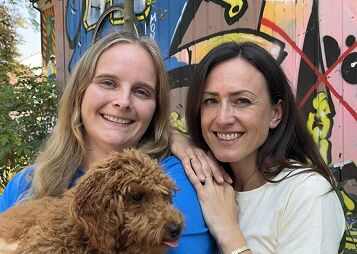 Zwei lächelnde Frauen, eine hält einen braunen Hund, bunte Graffiti im Hintergrund